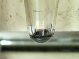 Close-up of a glass test tube with gray and black insolubles in the bottom