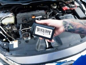 Tattooed arm holding a Blackstone oil sampling kit in front of an engine