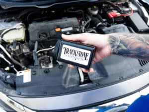 Tattooed arm holding a Blackstone oil sampling kit in front of an engine