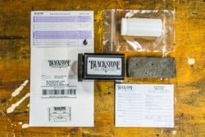 Disassembled kit on a workbench. The kit contains the outer black mailer and lid, a postage-paid return mailing envelope, gray absorbent material, a ziploc bag, an oil slip, instructions, and an oil change windshield reminder sticker.
