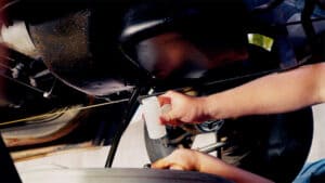 Hands holding a sampling bottle under an engine to catch oil as it drains out of the pan